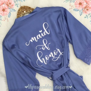 May include: A light blue satin robe with white lace trim on the sleeves. The back of the robe has the text "maid of honor" written in white cursive.