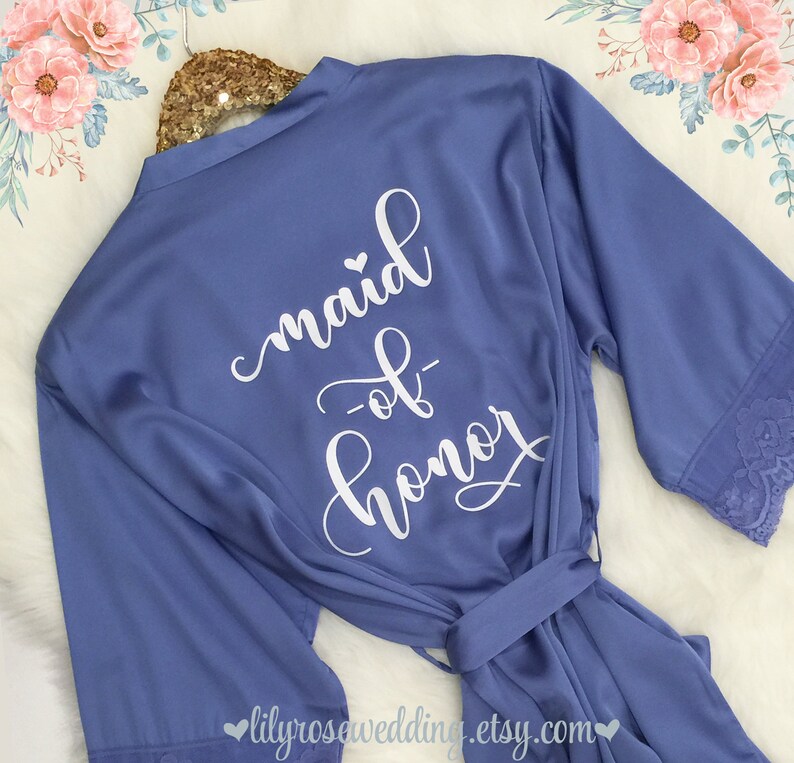 May include: A light blue satin robe with white lace trim on the sleeves. The back of the robe has the text "maid of honor" written in white cursive.
