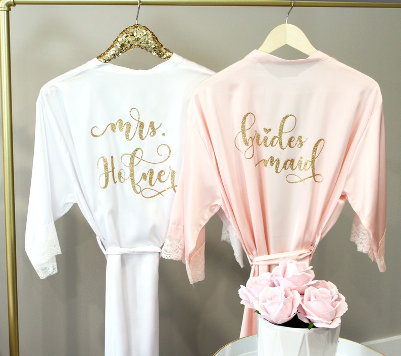 May include: Two satin robes with lace trim, one white with "mrs. Hofner" in gold glitter lettering and one pink with "bridesmaid" in gold glitter lettering.
