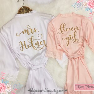 May include: Two satin robes with lace trim. The white robe has "mrs. Holner" written on the back in gold glitter. The pink robe has "flower girl" written on the back in gold glitter.