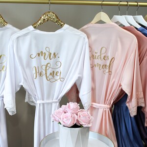 May include: Five white satin robes with gold lettering and lace trim. The robes are hanging on a gold rack. The robes say "bride", "mrs. Hofner", "bridesmaid", and two others are unreadable.