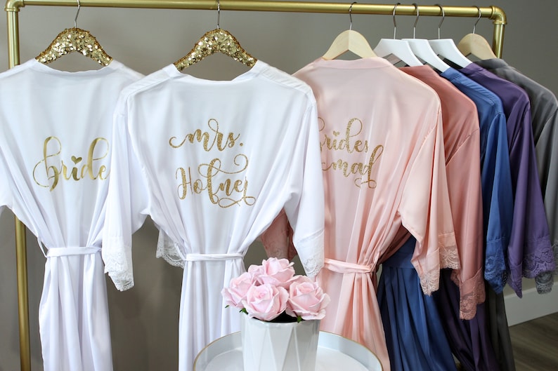 May include: Five white satin robes with gold lettering and lace trim. The robes are hanging on a gold rack. The robes say "bride", "mrs. Hofner", "bridesmaid", and two others are unreadable.