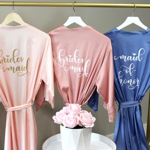 May include: Three satin robes hanging on a gold rack. The robes are in pink, light pink, and blue. The pink robes have the text "bridesmaid" in gold glitter and white lettering. The blue robe has the text "maid of honor" in white lettering.
