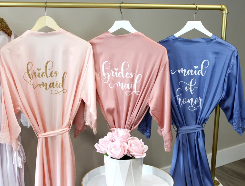 May include: Three satin robes hanging on a gold rack. The robes are in pink, light pink, and blue. The pink robes have the text "bridesmaid" in gold glitter and white lettering. The blue robe has the text "maid of honor" in white lettering.
