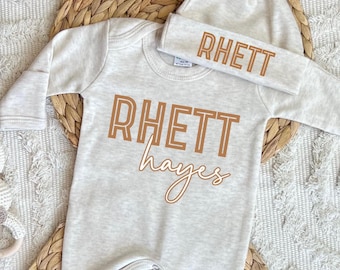 Könnte beinhalten: Beiges Baby-Set, bestehend aus einem Body und einer passenden Mütze. Der Body zeigt den Namen "RHETT" in großen, umrandeten braunen Buchstaben, darunter "hayes" in Schreibschrift. Auf der Mütze steht ebenfalls "RHETT" in derselben braunen Schrift.