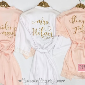 May include: Three satin robes with lace trim, one white and two blush pink, with gold glitter lettering. The white robe says "mrs. Holmer", one pink robe says "bridesmaid", and the other pink robe says "flower girl".