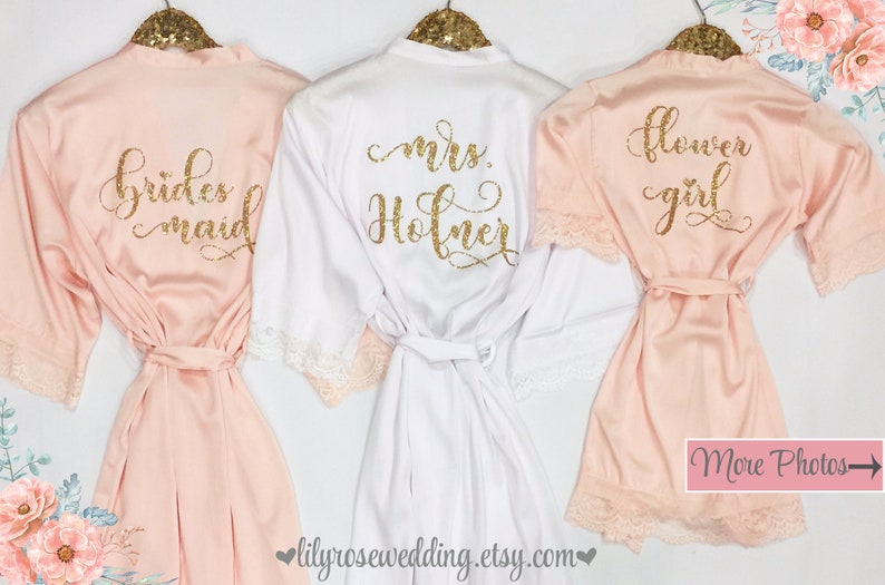 May include: Three satin robes with lace trim, one white and two blush pink, with gold glitter lettering. The white robe says "mrs. Holmer", one pink robe says "bridesmaid", and the other pink robe says "flower girl".