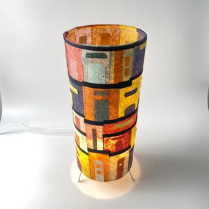 May include: A cylindrical lamp with a colorful, patchwork design. The lamp features a variety of rectangular shapes in shades of red, orange, yellow, purple, and green, separated by black bands. The lamp is lit from within, casting a warm glow.