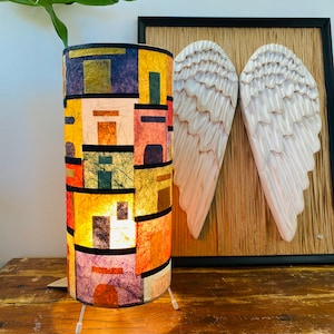 May include: A cylindrical table lamp with a colorful patchwork design. The lamp features a variety of rectangular shapes in shades of yellow, orange, purple, and blue. The lamp is on a wooden table, with a pair of carved wooden angel wings in the background.