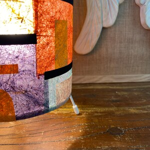May include: A cylindrical lamp with a colorful, textured lampshade. The shade features rectangular patches of orange, purple, and yellow, separated by black bands. A white cord extends from the base. The lamp sits on a wooden surface.