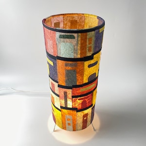 May include: A cylindrical table lamp with a colorful, geometric design. The lamp features a patchwork of various colors, including red, yellow, purple, and green, separated by black bands. The lamp is supported by three thin, white legs.