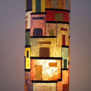 May include: A cylindrical table lamp with a colorful, geometric design. The lamp features a patchwork of rectangular and square shapes in various colors, including red, yellow, green, and purple. It stands on three small, white legs with wooden ball feet.