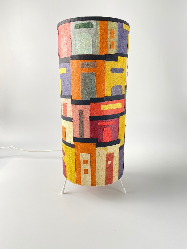 May include: A cylindrical lamp with a colorful, textured shade. The shade features a patchwork design of various rectangular shapes in shades of yellow, orange, purple, and red, separated by black bands. The lamp has a white, three-legged base.