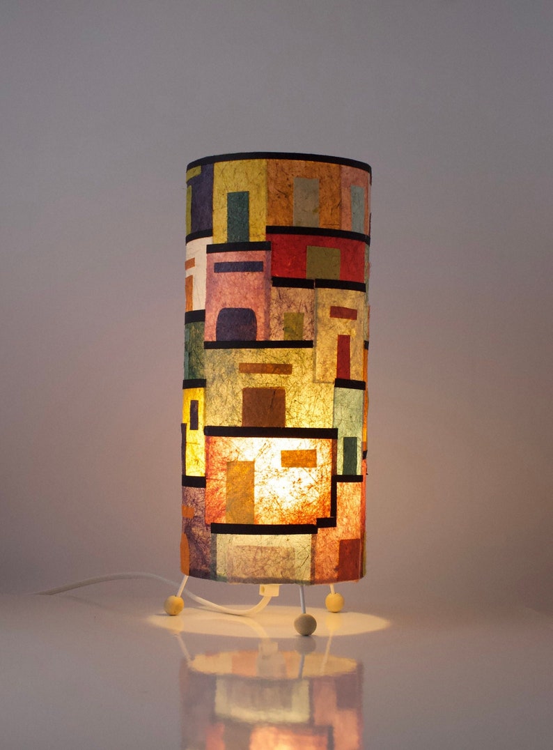 May include: A cylindrical table lamp with a colorful, geometric design. The lamp features a patchwork of rectangular shapes in various colors, including red, yellow, and blue, outlined in black. It stands on three small, white legs and has a white cord.
