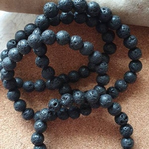 May include: Three strands of black lava rock bead bracelets. The beads are round and have a porous texture. The bracelets are displayed on a brown surface with a light-coloured stone in the background. Each bracelet is approximately 18 cm in circumference.