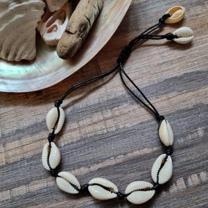 May include: A handcrafted bracelet made with white cowrie shells threaded onto a black cord. The shells are spaced and knotted, creating a beachy, bohemian style. The bracelet is displayed on a wooden surface with seashells in the background.