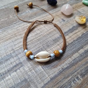 May include: A brown macrame bracelet with a white seashell centrepiece, wooden beads, and light blue beads. The adjustable bracelet is displayed on a wooden surface, with various gemstones in the background.