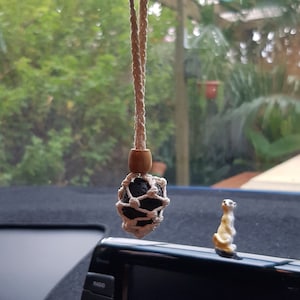 May include: A white string car air freshener with a black lava rock ball inside a white macrame net. The air freshener is hanging from the rear view mirror of a car.