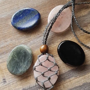 May include: A set of four smooth, polished stones, including a blue lapis lazuli, a grey labradorite, a black obsidian, and a pink rose quartz. The rose quartz is strung on a grey cord with a wooden bead and a macrame net.