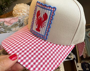 May include: A beige baseball cap featuring a red and white gingham brim and a rectangular patch on the front. The patch displays a red lobster illustration on a light blue striped background, framed by a red and white gingham border and blue scalloped trim.