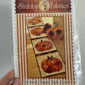 May include: A package for a patchwork pumpkin table runner. The table runner features three pumpkin designs in shades of orange and brown. The package has the words "Shabby Fabrics" and "Patchwork Pumpkin Table Runner".