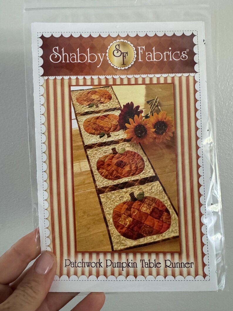 May include: A package for a patchwork pumpkin table runner. The table runner features three pumpkin designs in shades of orange and brown. The package has the words "Shabby Fabrics" and "Patchwork Pumpkin Table Runner".