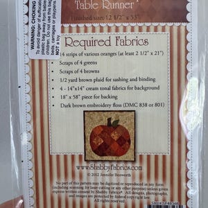 May include: A package for a "Patchwork Pumpkin Table Runner" with a finished size of 12 1/2" x 53". The package includes a list of required fabrics, including orange, green, and brown fabrics. A small image of a patchwork pumpkin is featured.