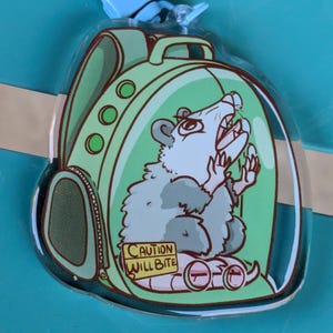 May include: A clear plastic keychain featuring a cartoon possum wearing a green backpack and glasses. The possum is inside a clear plastic bubble with a yellow warning sign that reads "CAUTION WILL BITE".