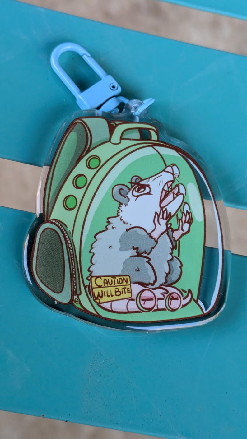 May include: A clear plastic keychain featuring a cartoon possum wearing a green backpack and glasses. The possum is inside a clear plastic bubble with a yellow warning sign that reads "CAUTION WILL BITE".