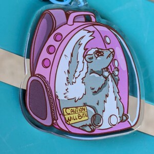 May include: A pink backpack charm featuring a cartoon skunk inside. The skunk is grey with a white stripe and tail. The backpack has a zipper and a warning label that reads "Caution Will Bite".