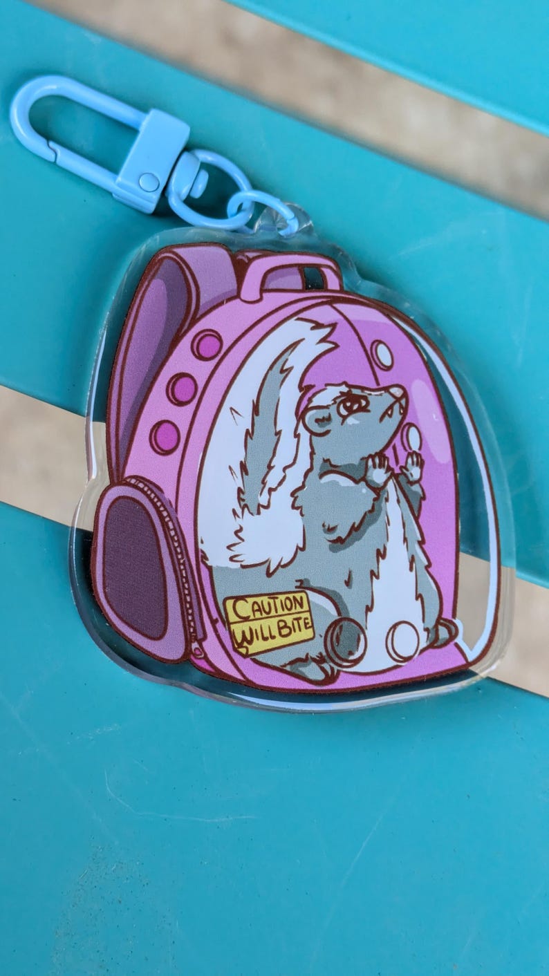 May include: A pink backpack charm featuring a cartoon skunk inside. The skunk is grey with a white stripe and tail. The backpack has a zipper and a warning label that reads "Caution Will Bite".