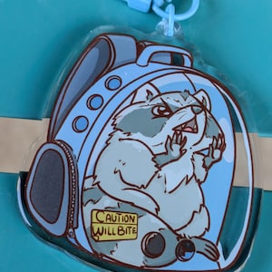 May include: A clear plastic keychain featuring a cartoon raccoon wearing a blue backpack. The raccoon is looking to the right and has a warning sign that reads "Caution Will Bite".