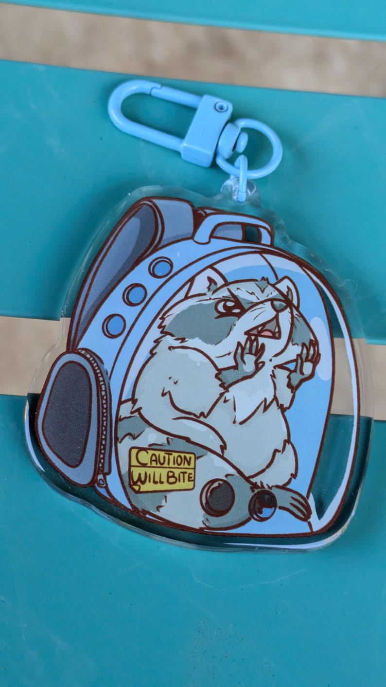 May include: A clear plastic keychain featuring a cartoon raccoon wearing a blue backpack. The raccoon is looking to the right and has a warning sign that reads "Caution Will Bite".