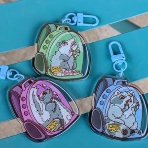 May include: Three colourful keychains featuring cartoon animals in a backpack-shaped bubble. Each keychain has a blue, green, or pink backpack with a different animal inside: a raccoon, opossum, and skunk. The text "Caution Will Bite" is on each keychain.