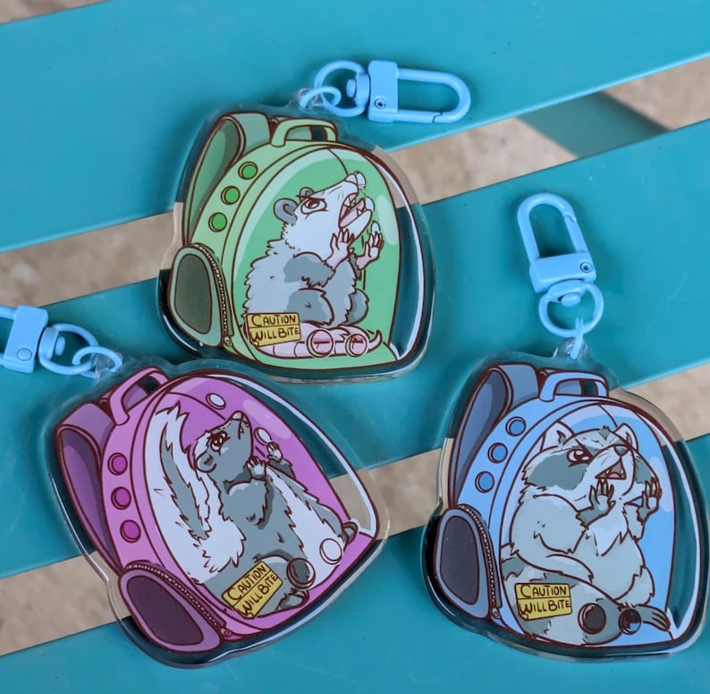 May include: Three colourful keychains featuring cartoon animals in a backpack-shaped bubble. Each keychain has a blue, green, or pink backpack with a different animal inside: a raccoon, opossum, and skunk. The text "Caution Will Bite" is on each keychain.