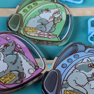 May include: Three colourful keychains featuring cartoon animals in a backpack-shaped bubble. The keychains are made of clear acrylic and feature a blue raccoon, a pink skunk, and a green opossum. Each keychain has a warning label that reads "Caution Will Bite".