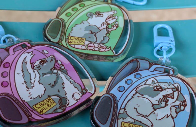 May include: Three colourful keychains featuring cartoon animals in a backpack-shaped bubble. The keychains are made of clear acrylic and feature a blue raccoon, a pink skunk, and a green opossum. Each keychain has a warning label that reads "Caution Will Bite".