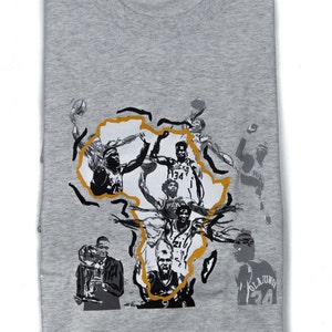 Può includere: Maglietta grigia con una stampa grafica di un contorno del continente africano in oro. La grafica presenta diversi giocatori di basket in bianco e nero, tra cui Hakeem Olajuwon, con il testo "Olajuwon 34" in basso a destra.