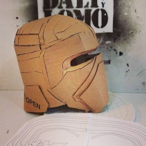 May include: A cardboard helmet with a visor, partially assembled, with the word "OPEN" printed on the side. The helmet is sitting on a white surface next to a sheet of paper with a black line drawing of a helmet.