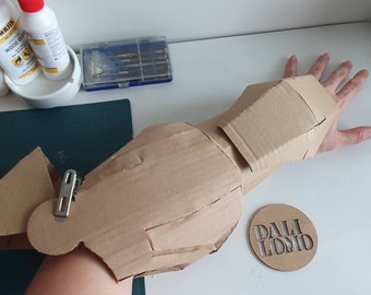 May include: A cardboard arm piece for a costume, with a metal clip attached. The arm piece is made of multiple pieces of cardboard, and is brown in color. The arm piece is on a person's arm, and the person's hand is visible in the image. A wooden circle with the text "DALI LOMO" is visible in the lower right corner of the image.