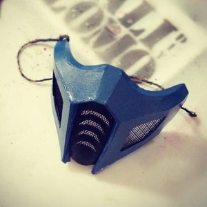 Puede incluir: Una máscara de plástico azul y negro con una malla que cubre los ojos. La máscara tiene un diseño en relieve y está atada con una cuerda.