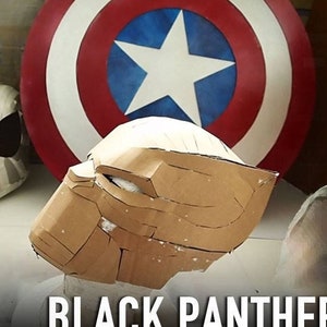 May include: A brown cardboard Black Panther mask is being made. The mask is sitting on a white surface in front of a red, white, and blue Captain America shield.