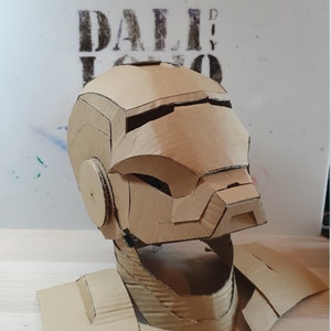 May include: A cardboard Iron Man helmet with a brown and tan color scheme. The helmet is made of cardboard and has a detailed design.