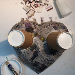 May include: A heart-shaped papier-m&acirc;ch&eacute; sculpture covered in newspaper clippings. Two brown paper cups with white tops are attached to the sculpture.
