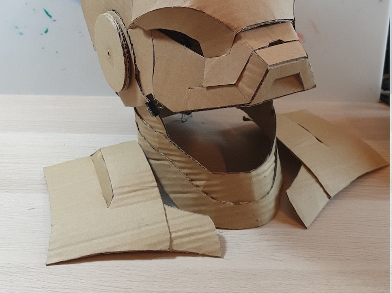 May include: A cardboard helmet with a face mask and neck piece. The helmet is made of brown cardboard and has a textured surface. The helmet is in pieces and is ready to be assembled.