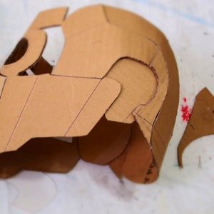 May include: A cardboard helmet is being assembled with a hot glue gun. The helmet is brown and has a design drawn on it. The helmet is being assembled on a white surface.