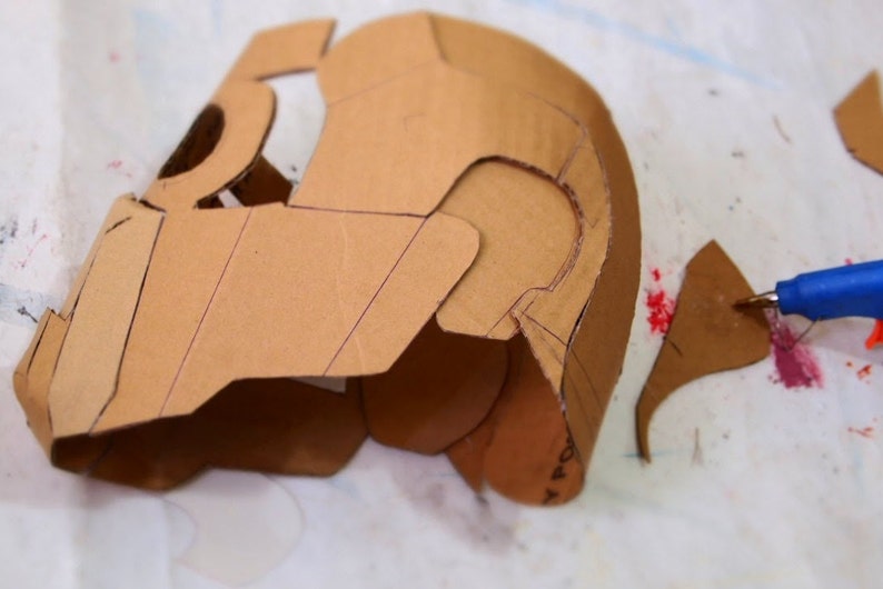May include: A cardboard helmet is being assembled with a hot glue gun. The helmet is brown and has a design drawn on it. The helmet is being assembled on a white surface.