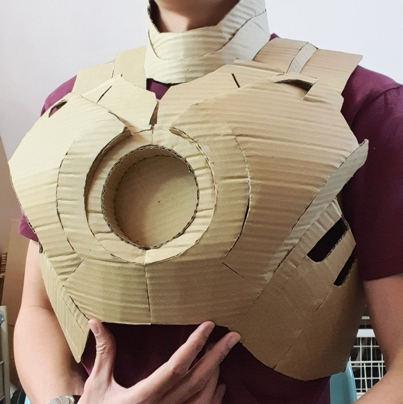Op de afbeelding: Een bruin kartonnen borstpantser met een cirkelvormig ontwerp in het midden. Het pantser heeft een gelaagde constructie met een bijpassende kraag. De persoon draagt een bordeauxrood shirt.