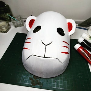 Peut inclure: Un masque blanc avec des d&eacute;tails rouges et noirs. Le masque a un visage de chat avec un petit nez et de grands yeux. Le masque est sur un tapis de coupe vert.