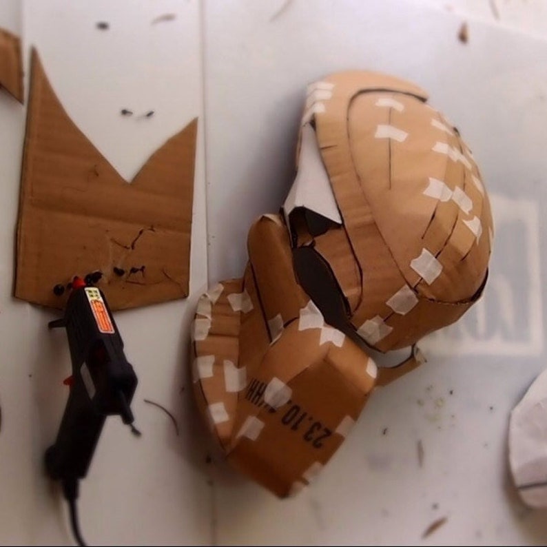 May include: A cardboard helmet is being assembled with tape and glue. The helmet is brown and has a white label that says "23 TO 10".